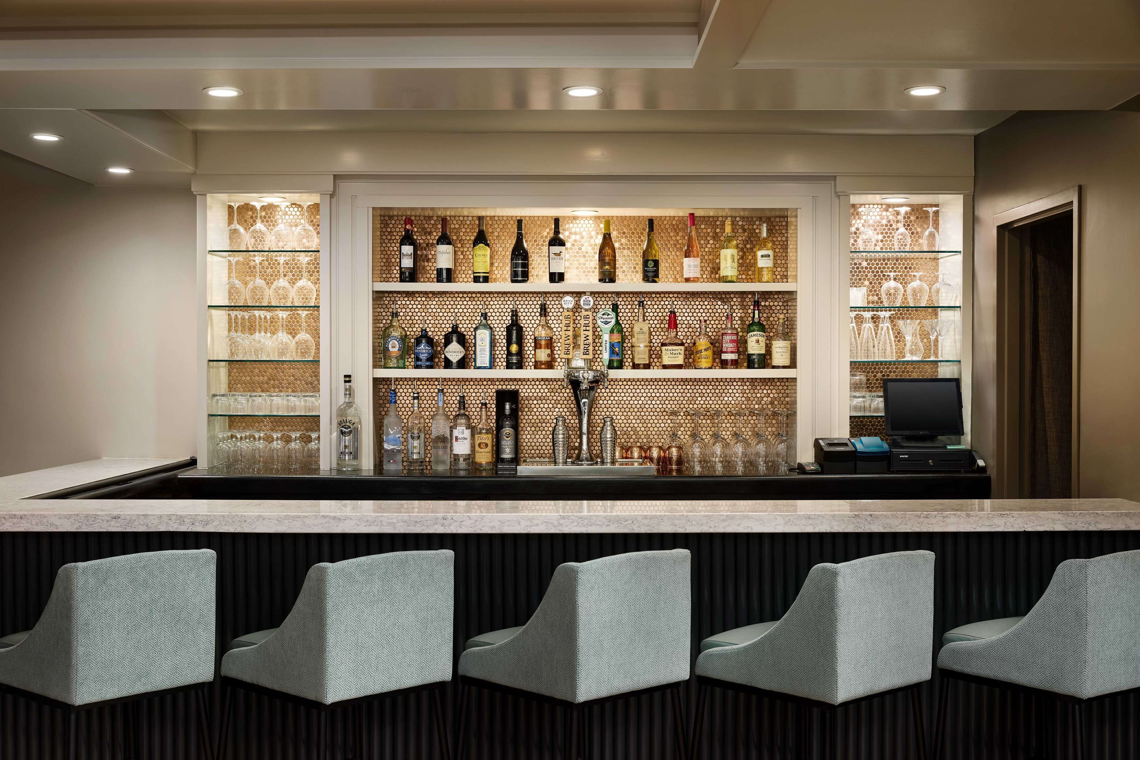 A bar area with a long counter, multiple shelves of bottles, and a computer. There are four upholstered bar stools in front.