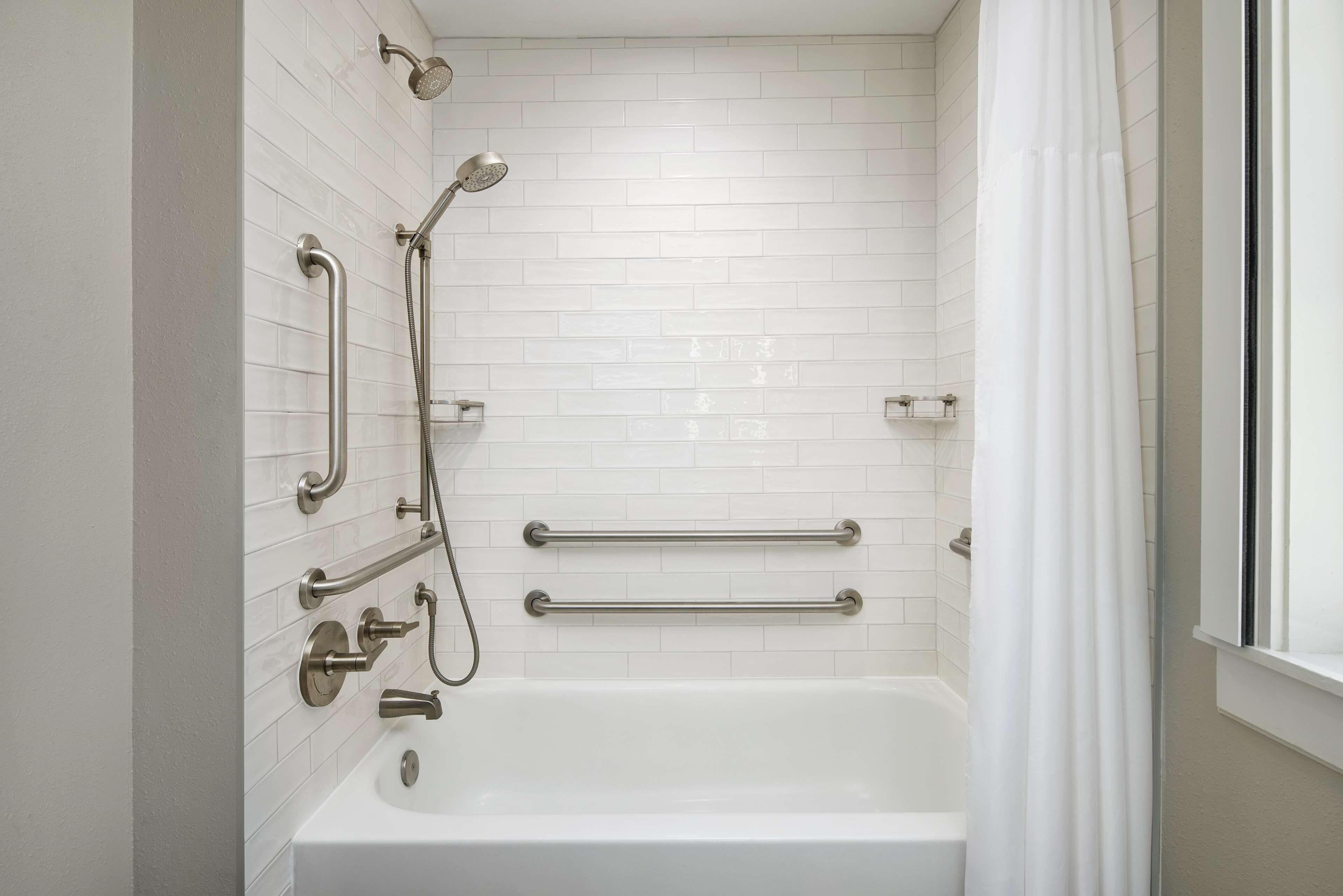 A modern bathroom with white tiled walls, a white bathtub, and grab bars for accessibility.