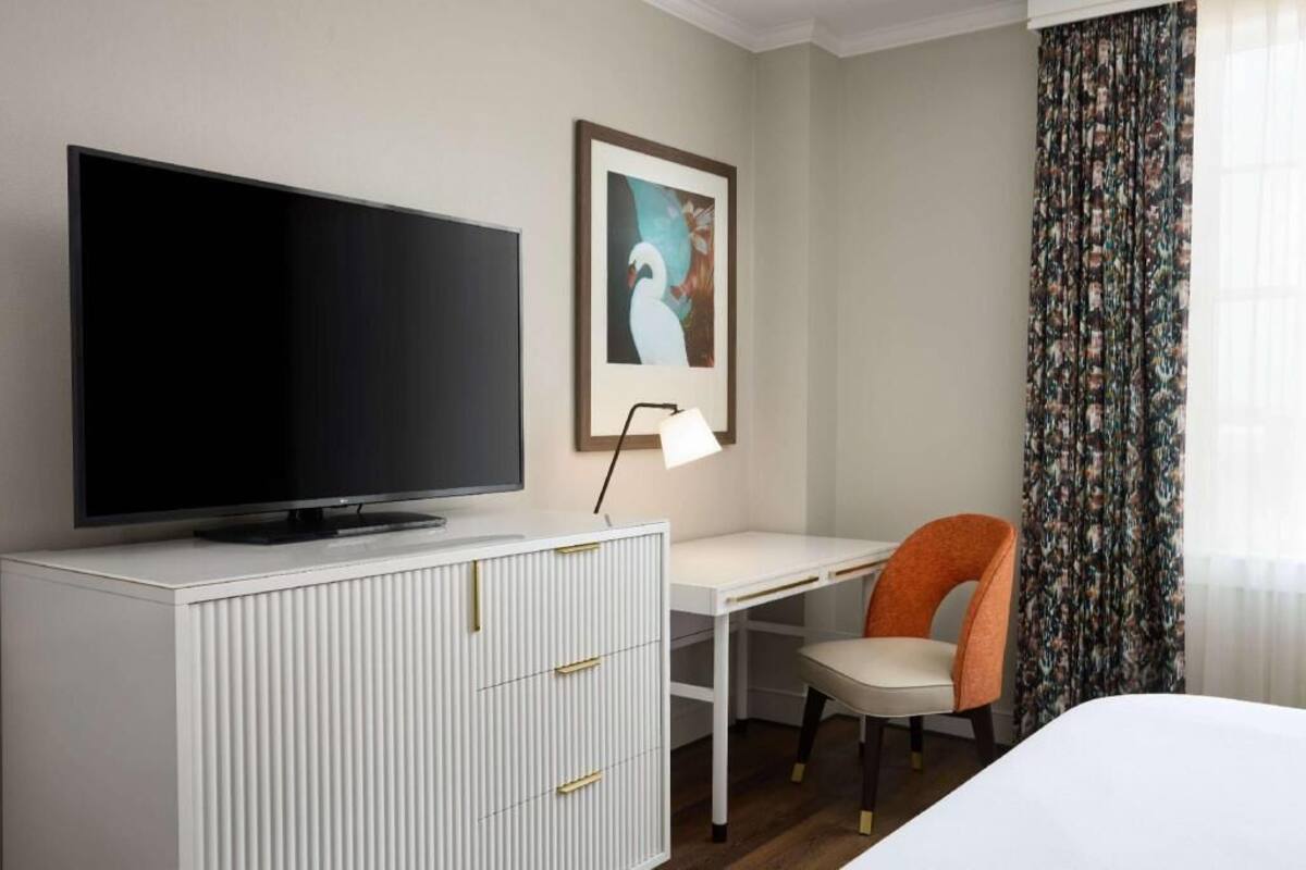 Habitación de hotel con televisión de pantalla plana sobre un mueble blanco, un escritorio con una silla, un cuadro en la pared y una ventana con cortinas.