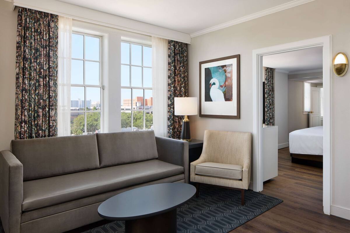 Habitación de hotel con sofá, sillón y una mesita. Se ve una cama al fondo y una ventana con cortinas.