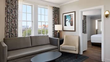 A hotel room with a sofa, armchair, and a small table. There is a bed in the background and a window with curtains.