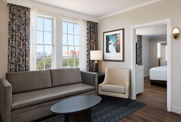 A hotel room with a sofa, armchair, and a small table. There is a bed in the background and a window with curtains. - The Terrace Hotel Lakeland, Tapestry Collection by Hilton (Lakeland)