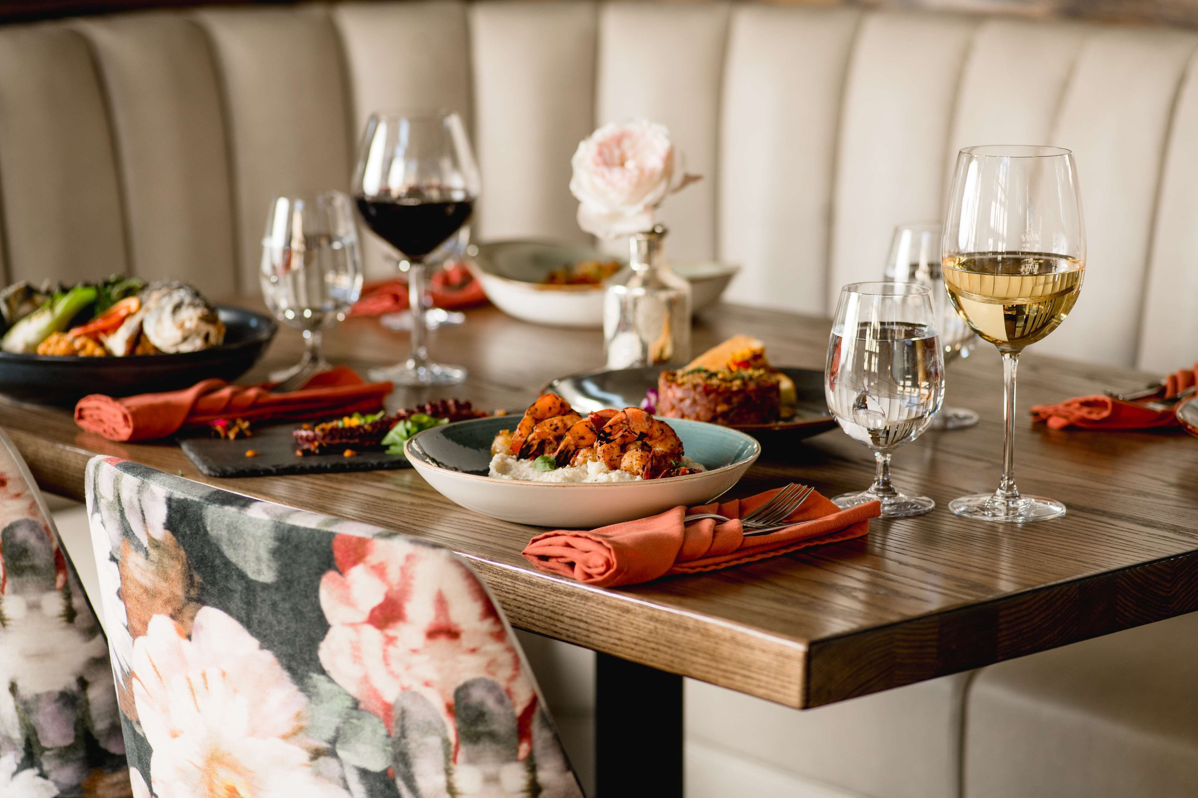 A dining table set with plates of food, wine glasses, and a rose in a vase.