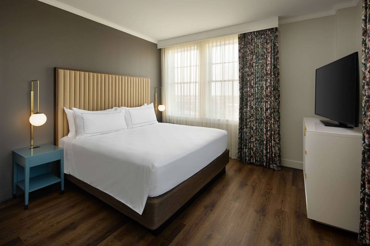 Habitación de hotel con una cama grande, una mesita de noche con lámpara, un televisor y una ventana con cortinas.