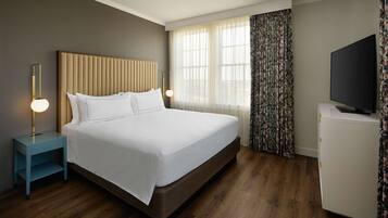 Habitación de hotel con una cama grande, una mesita de noche con lámpara, un televisor y una ventana con cortinas.