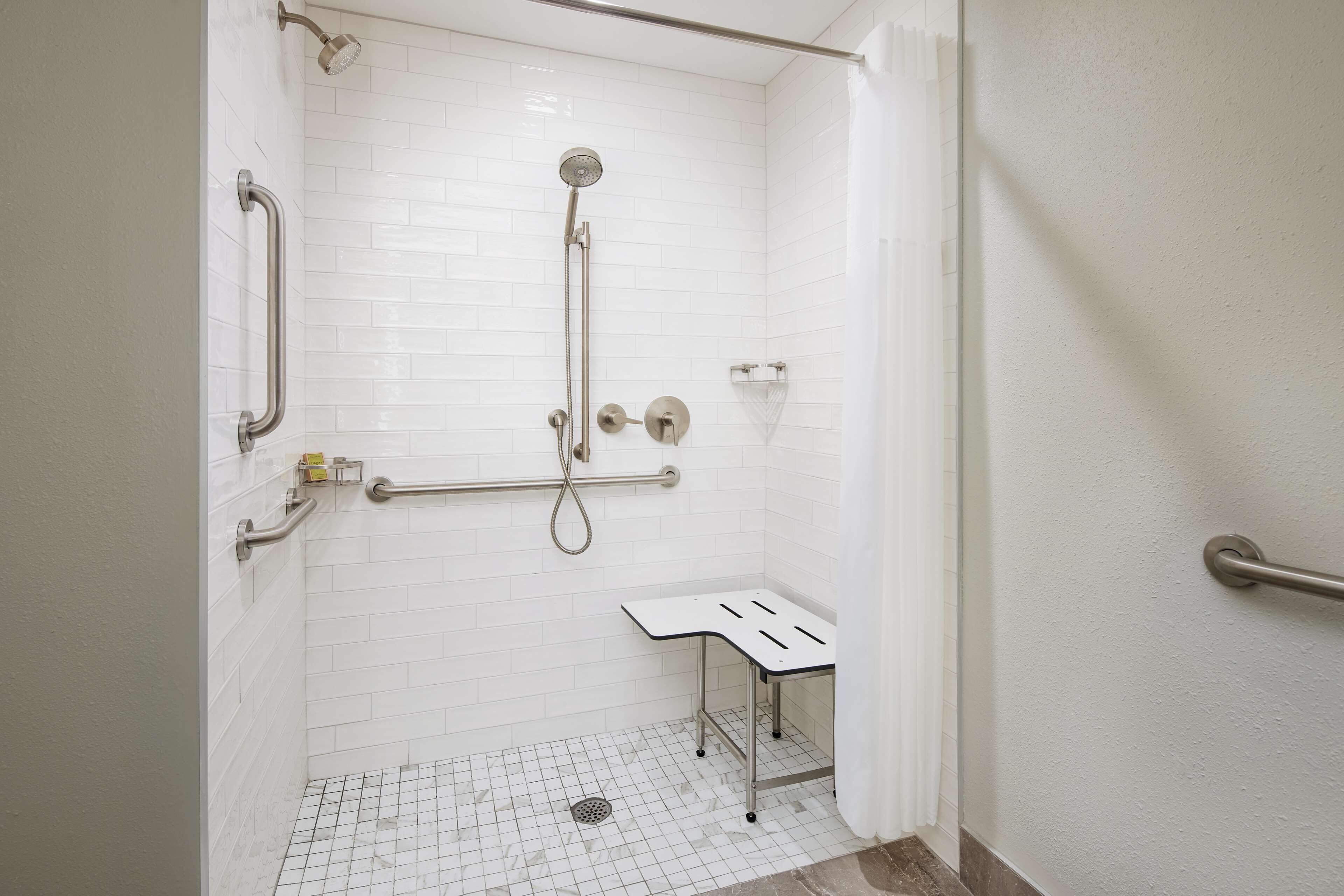 A white-tiled shower with a handheld showerhead, grab bars, and a small table.