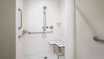 A white-tiled shower with a handheld showerhead, grab bars, and a small table.