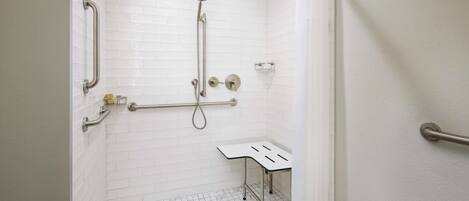 A white-tiled shower with a handheld showerhead, grab bars, and a small table.