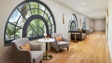 Un vestíbulo de hotel con amplias ventanas arqueadas, un área de descanso con sillones y una mesa redonda, y piso de madera.