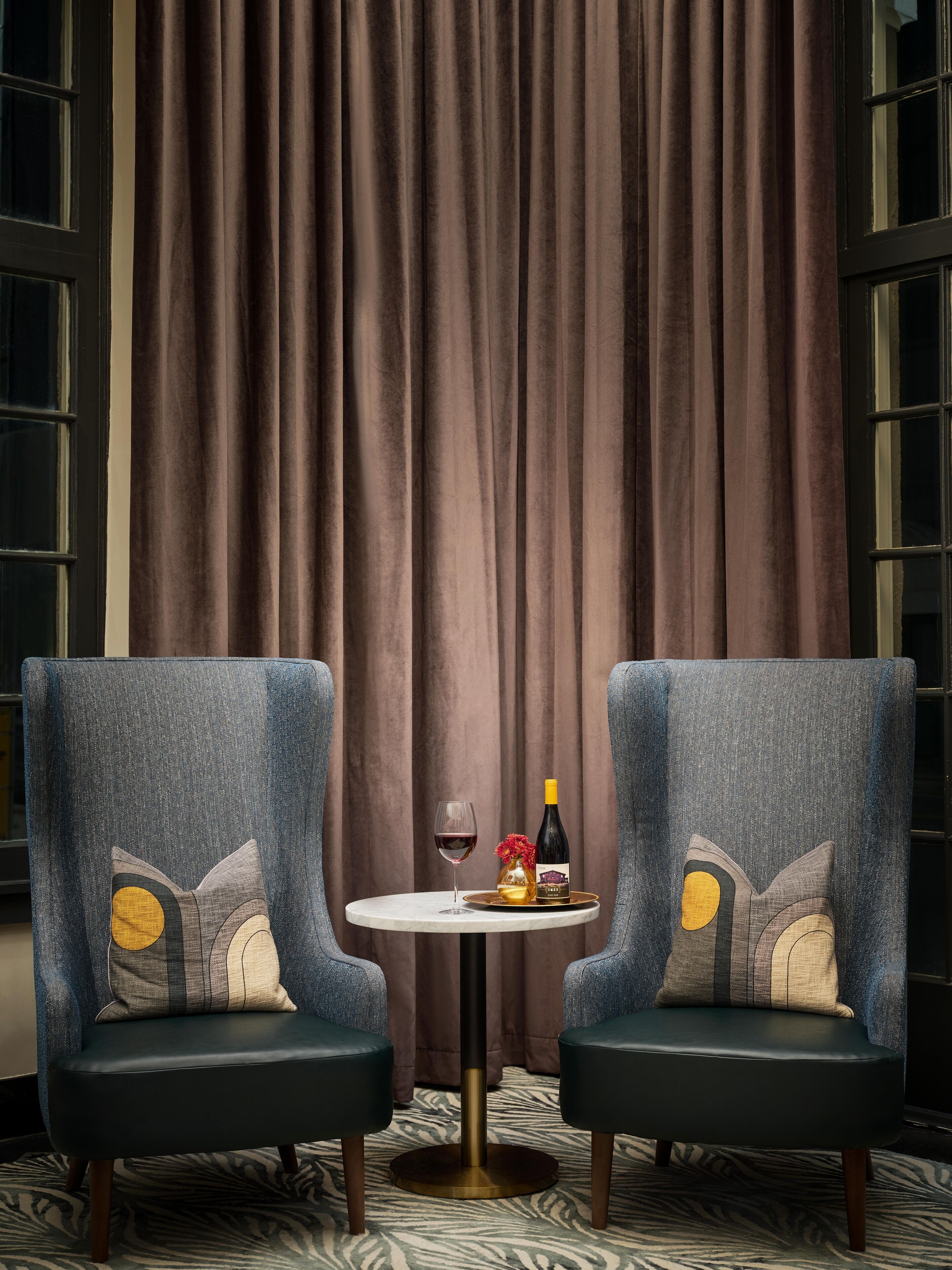 A room with two upholstered armchairs, a small round table, a bottle of wine, and a glass.