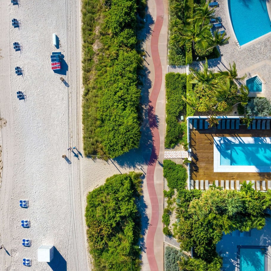 An aerial view of a beachside resort with a swimming pool, beach chairs, and a pathway.