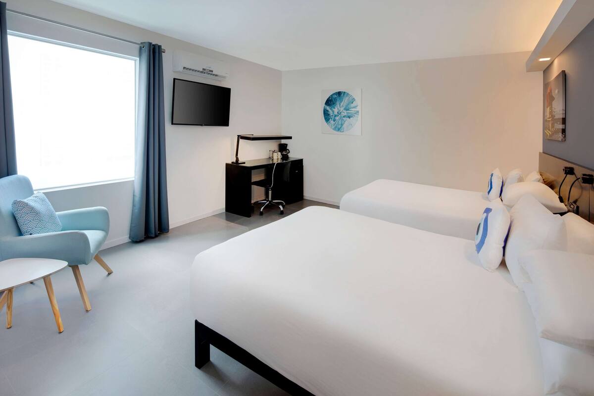 A modern hotel room with a large bed, a desk with a chair, a flat-screen TV, and a window with curtains.