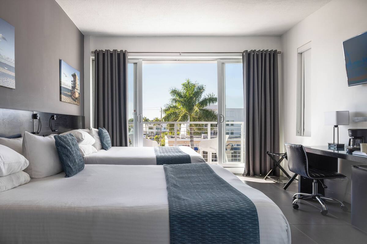 A hotel room with two beds, a desk, and a view of palm trees and buildings.