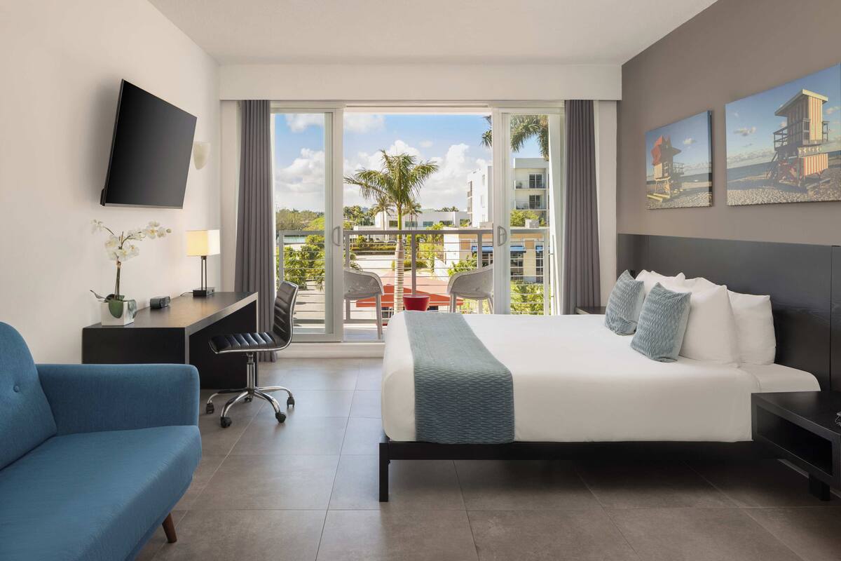 A hotel room with a bed, a desk, a chair, a TV, and a balcony with a view of palm trees and buildings.