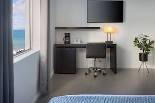 Habitación de hotel con cama, escritorio con silla, televisor, cafetera, planta y vista al océano.