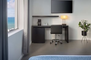 Habitación de hotel con cama, escritorio con silla, televisor, cafetera, planta y vista al océano.