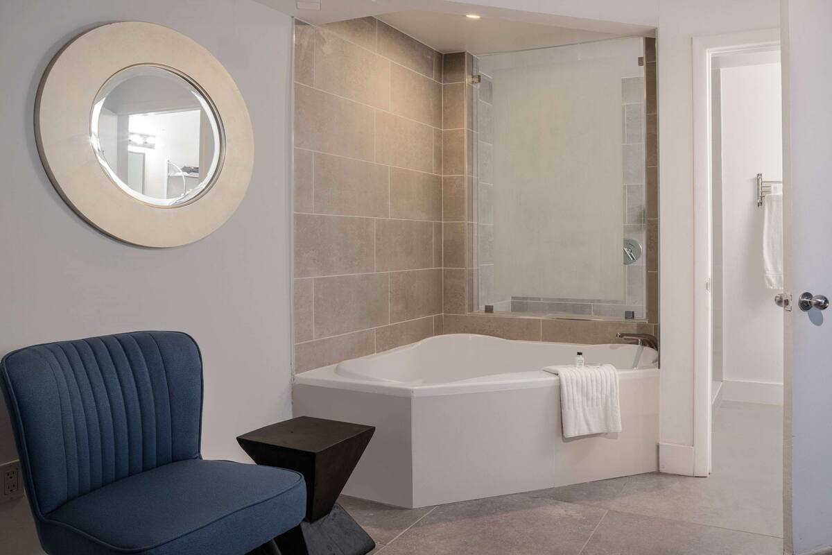 A modern bathroom with a bathtub, a round mirror, and a blue chair.