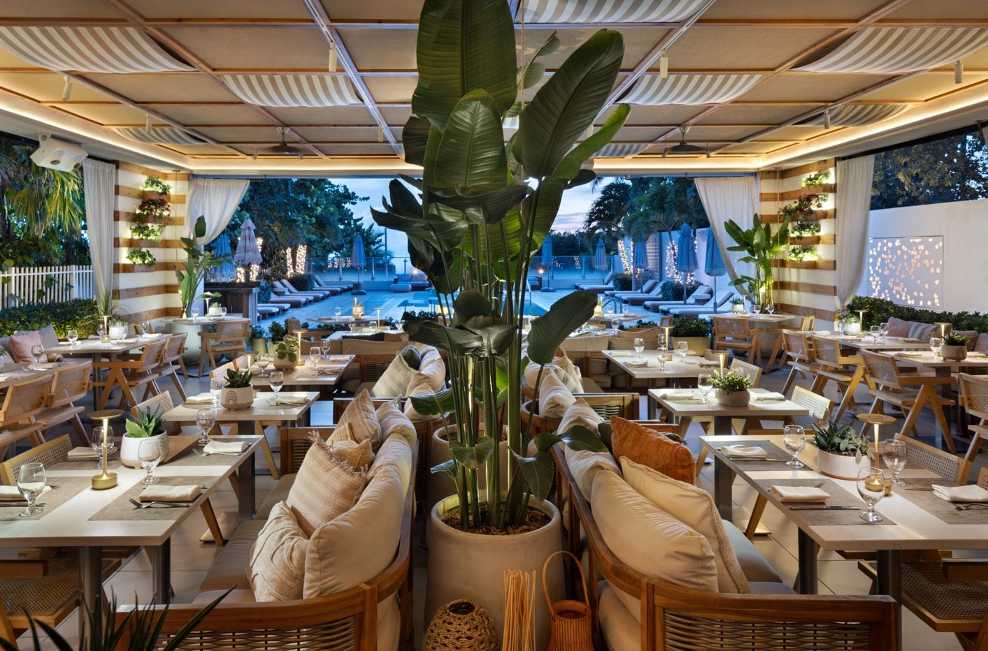 Un restaurante con varias mesas y sillas, plantas en macetas y vista a una piscina.