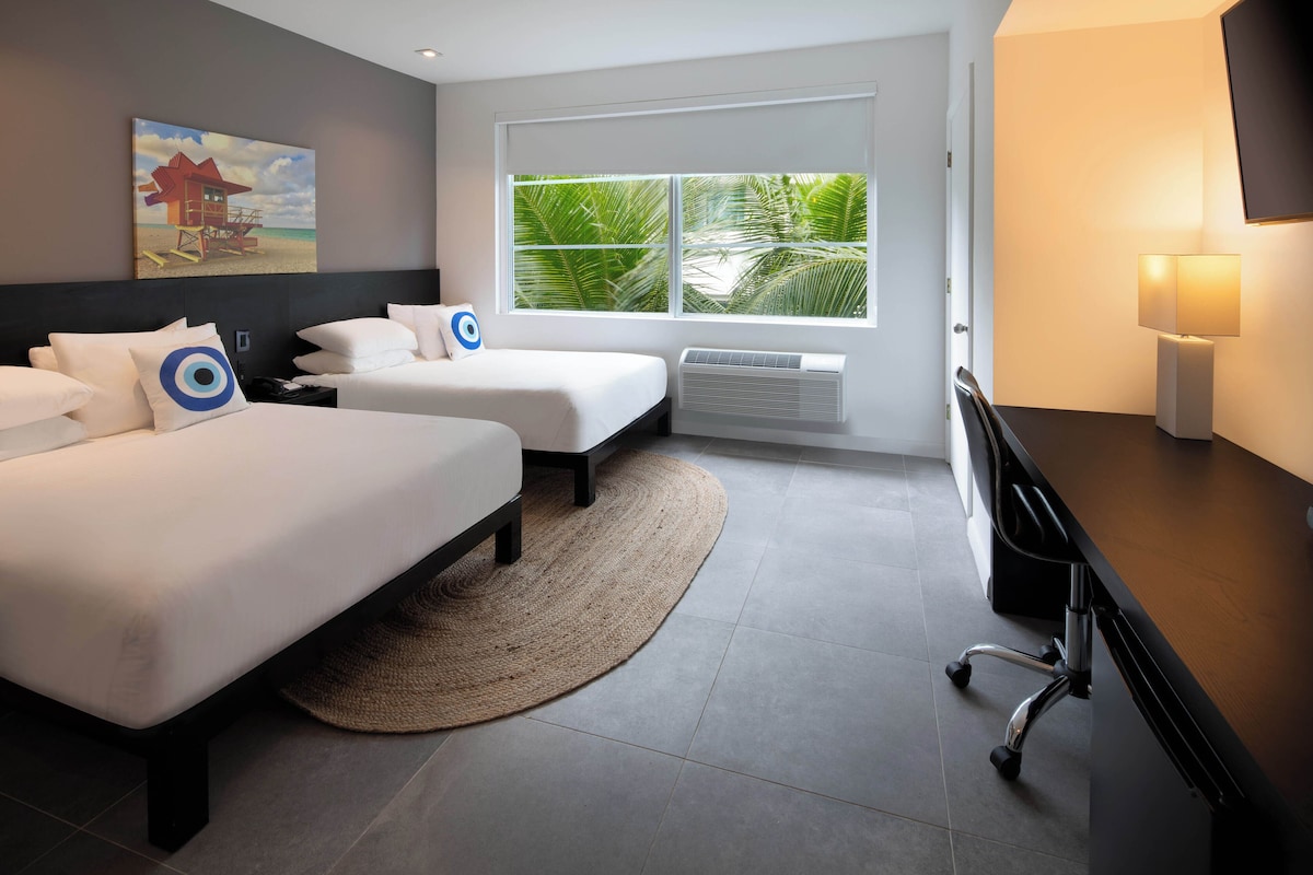 Una habitación de hotel moderna con dos camas, un escritorio, una silla, un ventanal con vistas a palmeras y un cuadro en la pared.
