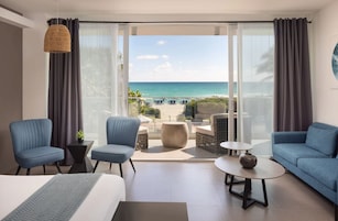 Habitación de hotel con cama, dos sillones, un sofá, una mesita y vista a la playa.