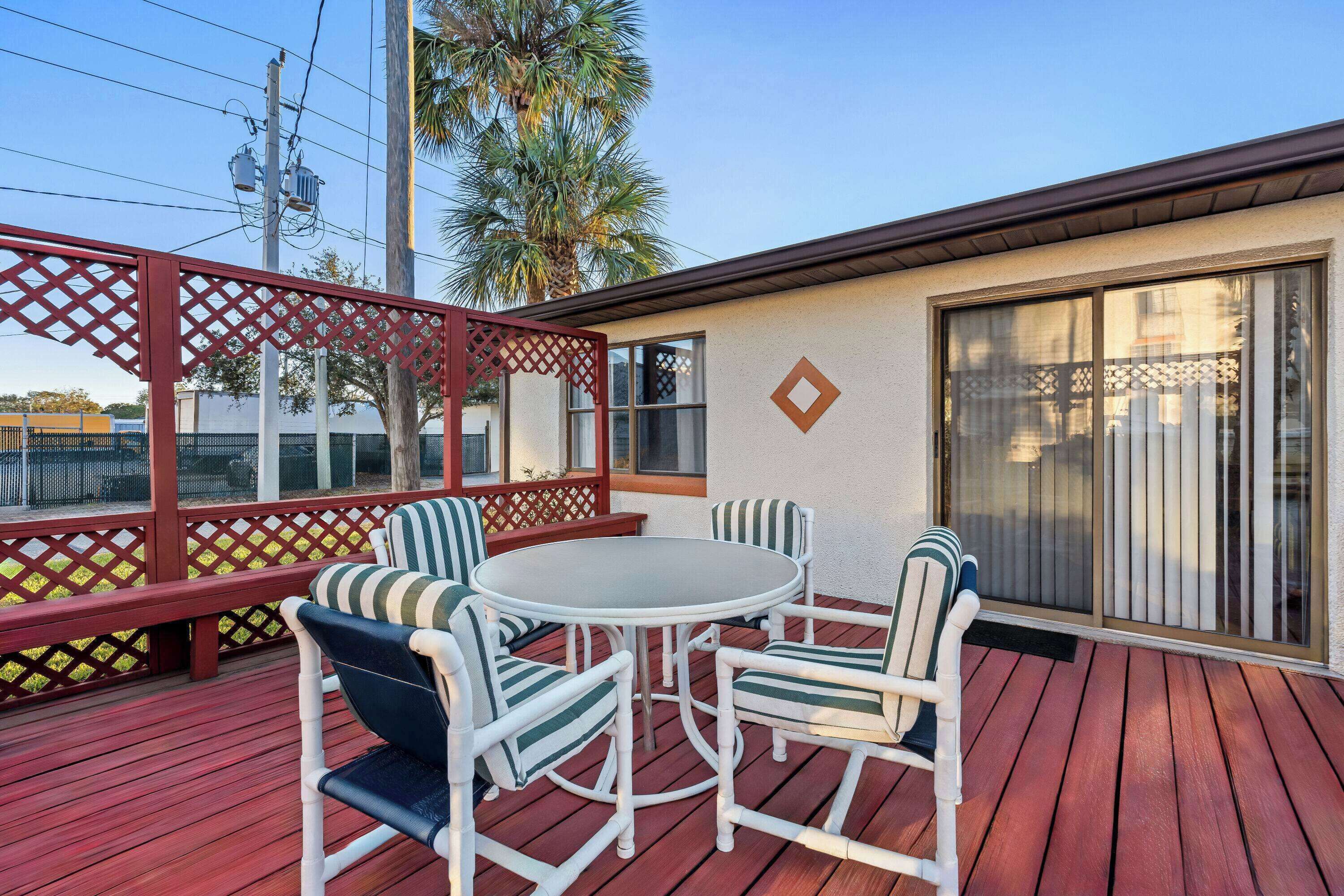 A covered outdoor patio with a round table and four chairs, a wooden deck, and a building with a large window and a door.