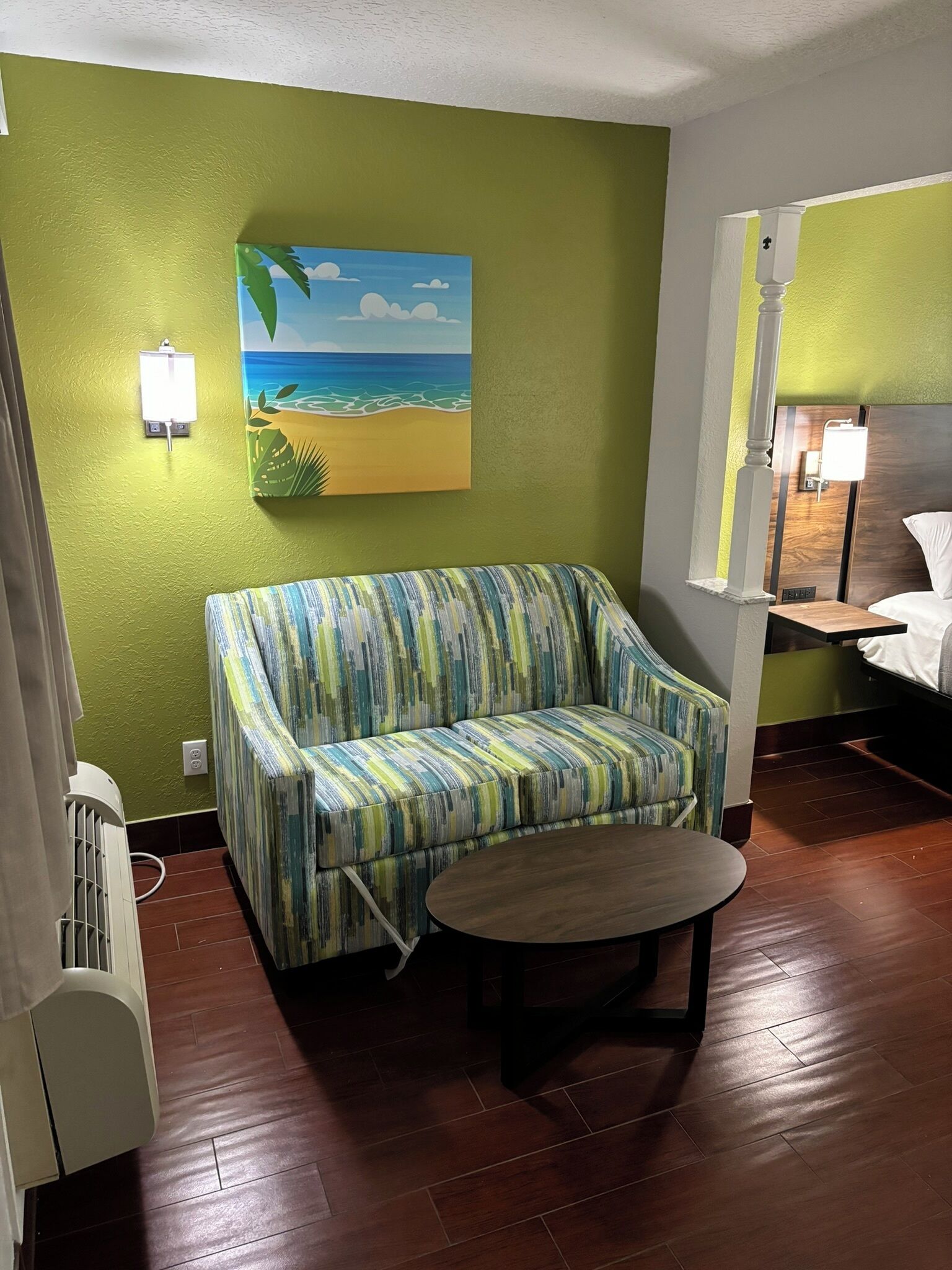 A room with a green wall, a painting of a beach, a striped sofa, a round table, and a wooden floor.