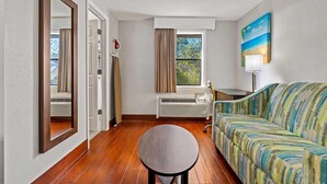 Una habitación de hotel con piso de madera, un sofá, una mesa redonda, una ventana con cortinas, un radiador y un cuadro en la pared.