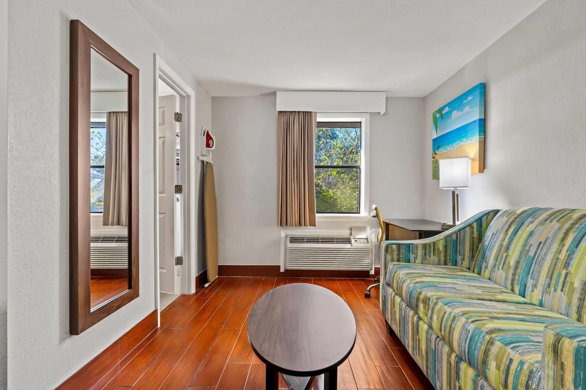 Habitació d'hotel amb sòl de fusta, un sofà, una taula rodona, una finestra amb cortines, un radiador i un quadre a la paret.