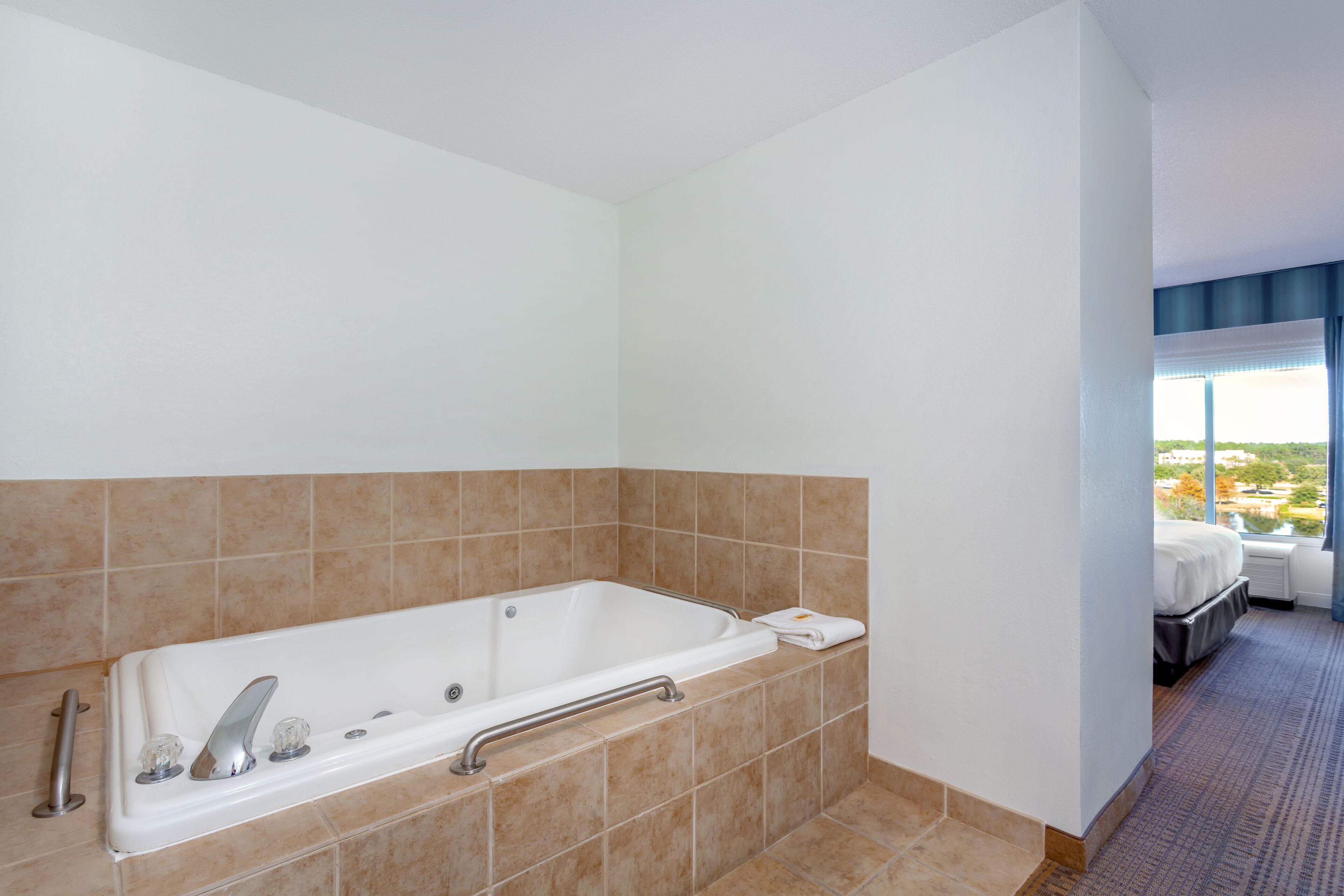 A modern bathroom with a large bathtub, a handrail, and a view of the city through a window.