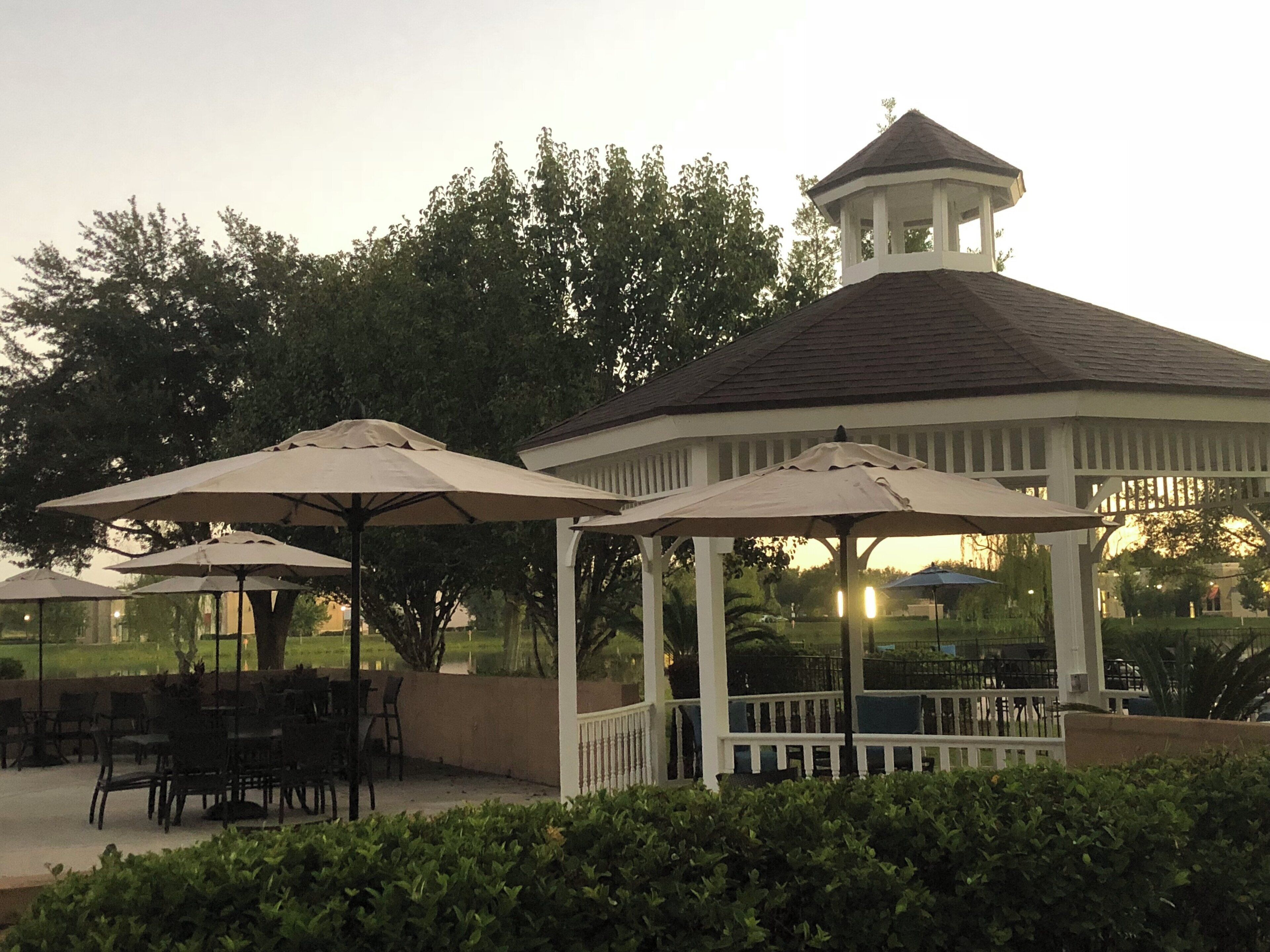 A gazebo with a white exterior and a peaked roof, surrounded by greenery and outdoor seating.