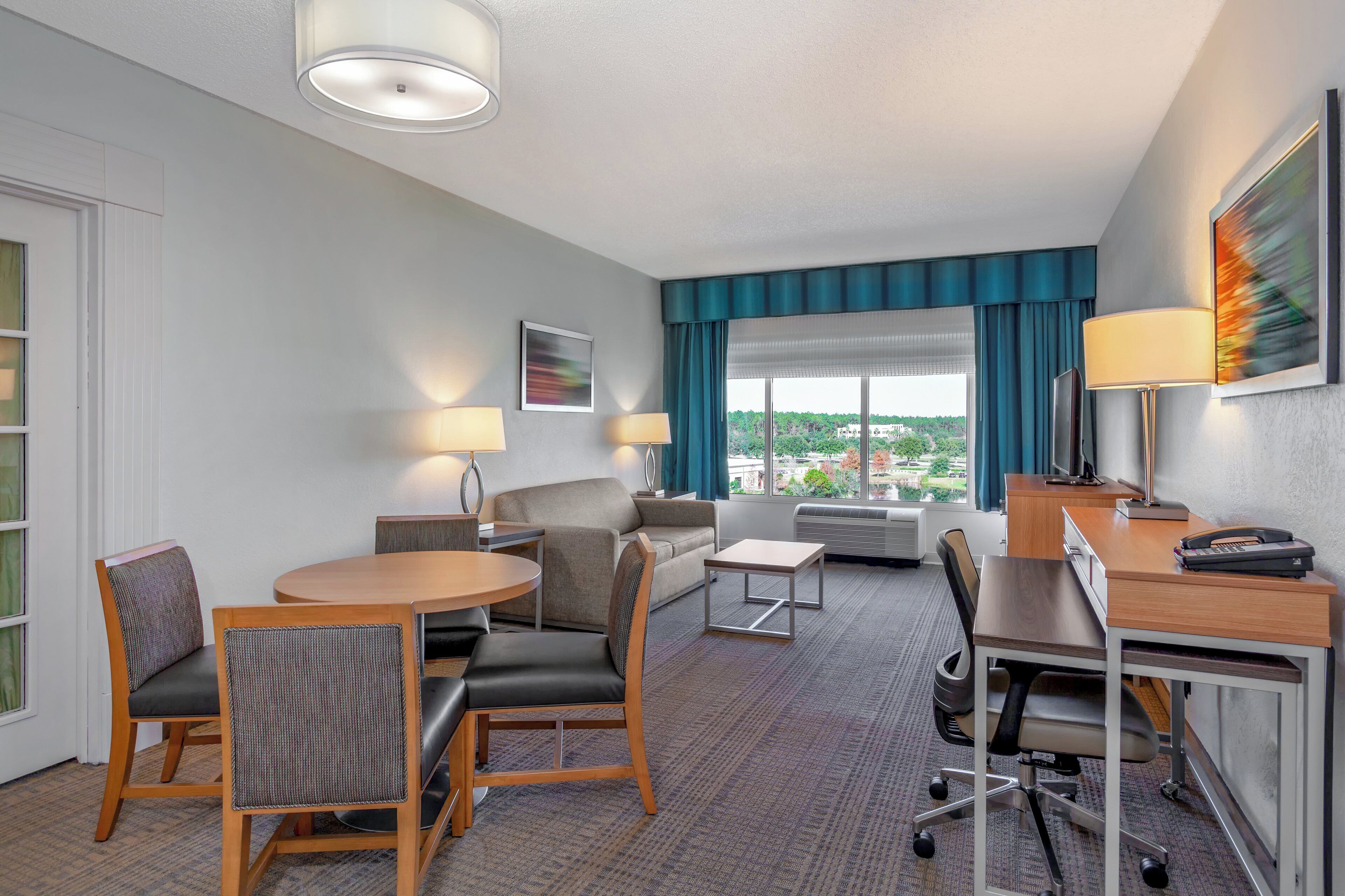 A hotel room with a dining area, a sofa, a desk with a telephone, and a large window with a view.