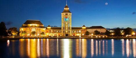 Un hotel frente al agua con una imponente torre de reloj, reflejos de luces sobre el agua.