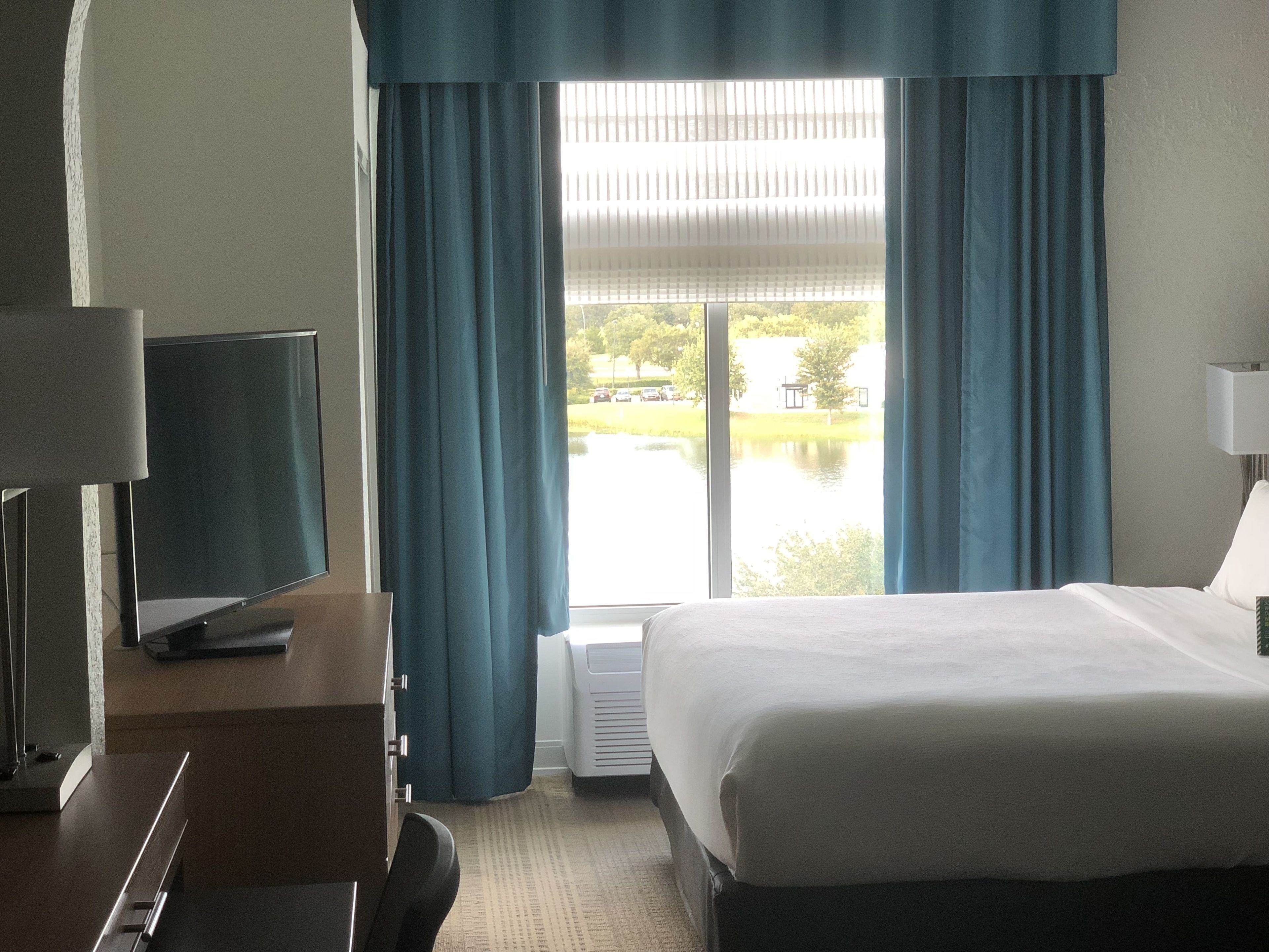 Ein Hotelzimmer mit Bett, Fernseher auf einem Schrank, Schreibtisch und einem Fenster mit Vorhängen.