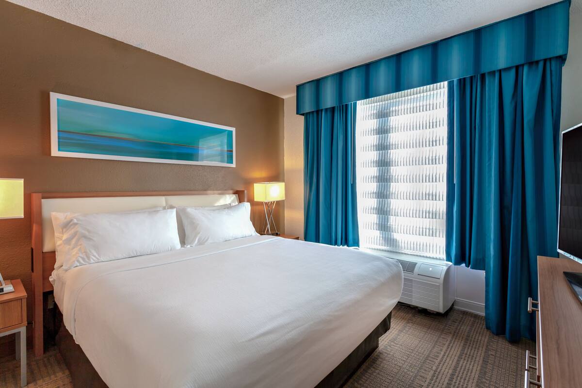 Une chambre d’hôtel avec un grand lit, un téléviseur à écran plat, une fenêtre avec des rideaux bleus et un tableau au mur.