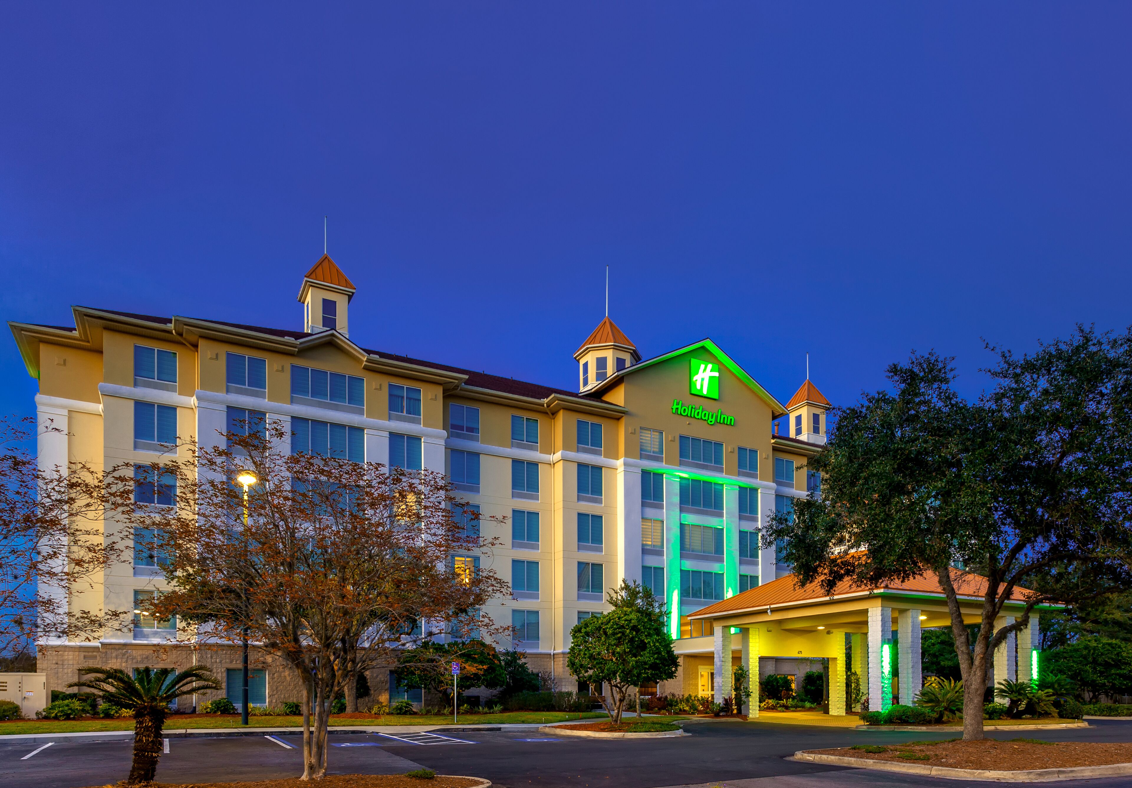 Um hotel Holiday Inn com um letreiro verde grande, várias janelas e uma entrada bem iluminada.