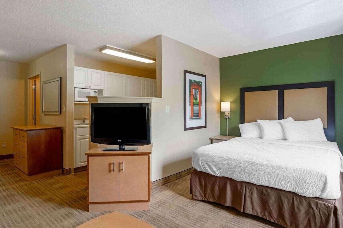 A hotel room with a bed, a TV on a stand, a kitchenette, and a bathroom.