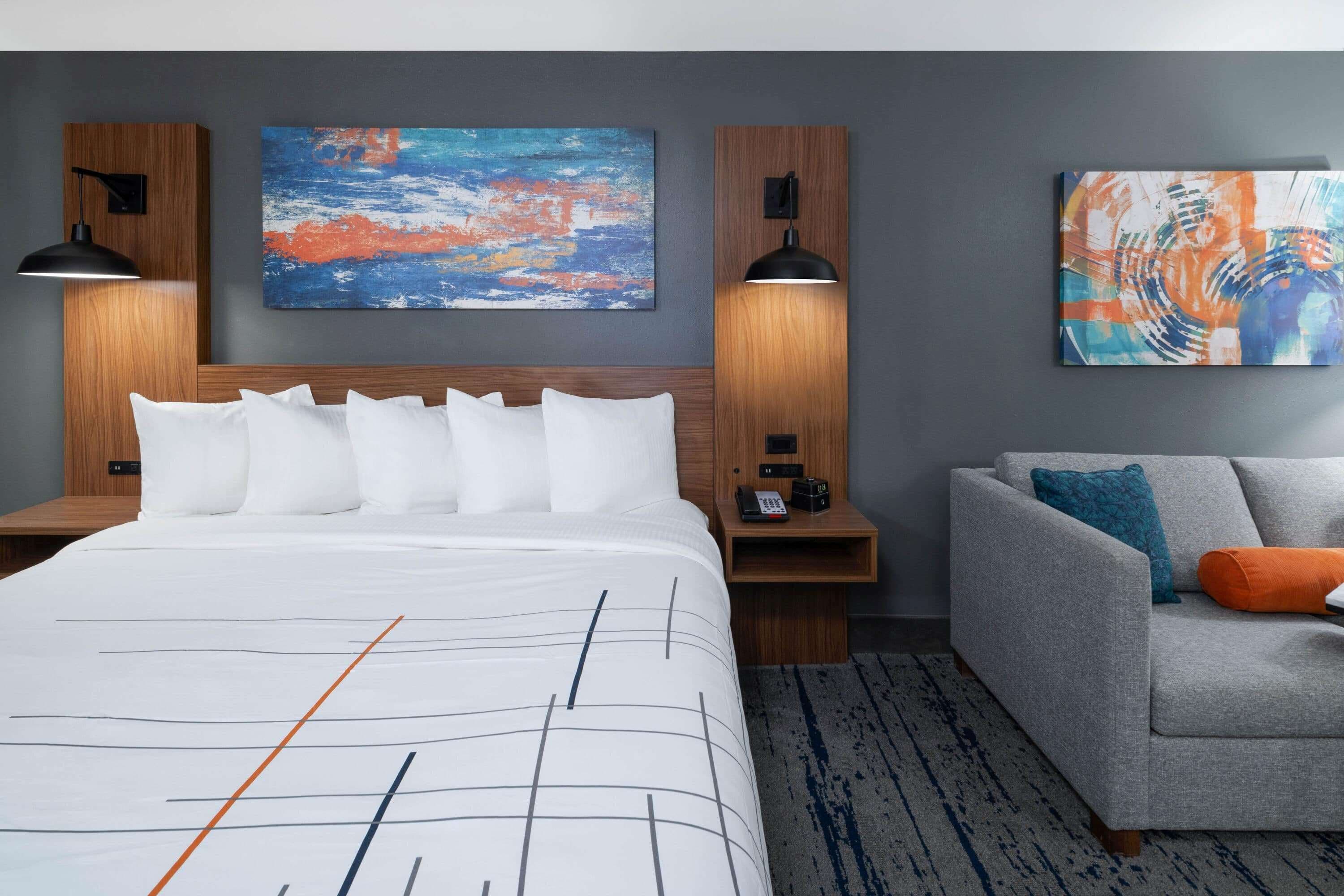A hotel room with a large bed, a grey sofa, two wall-mounted lamps, and two abstract paintings.