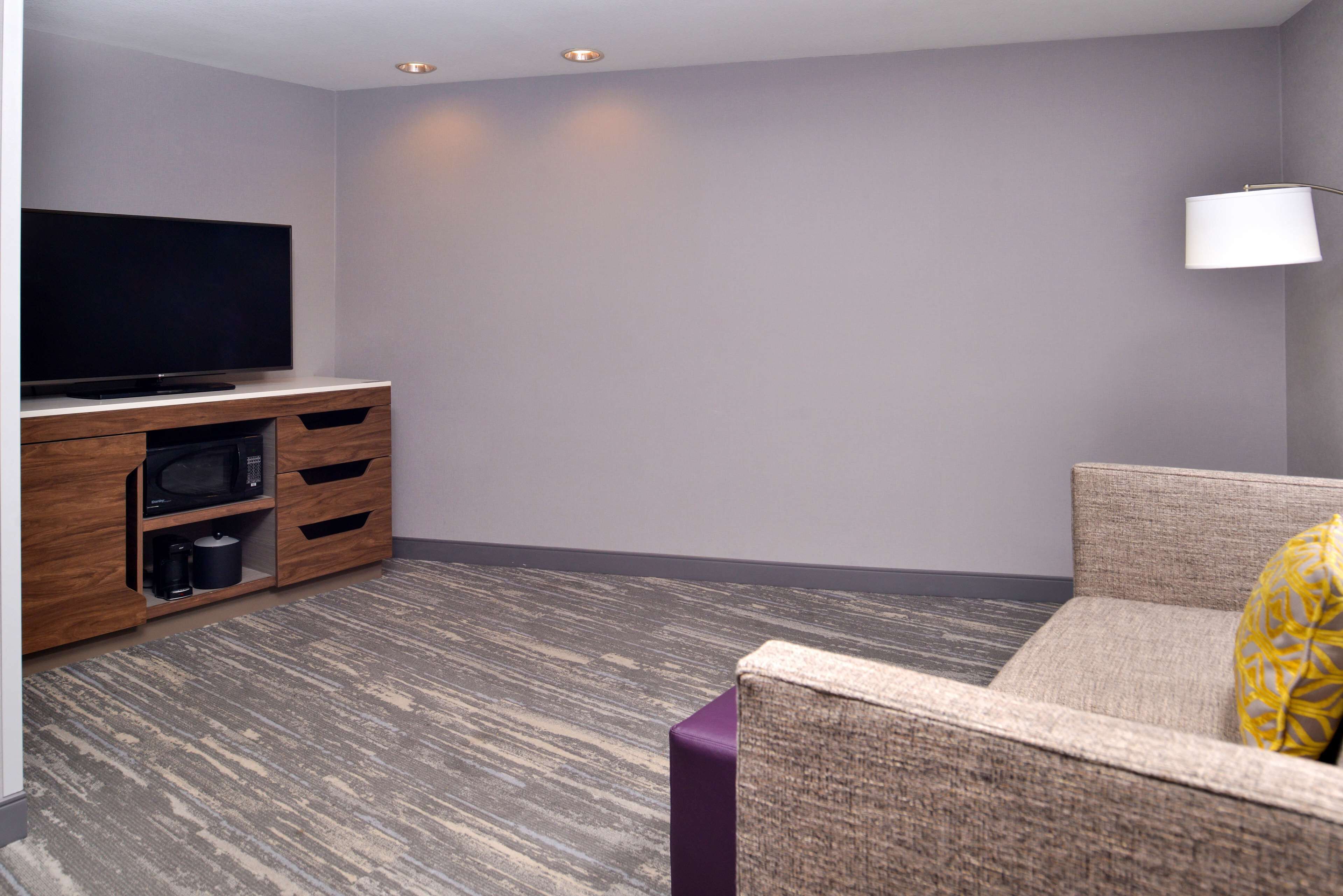 A modern living room with a flat-screen TV on a wooden cabinet, a sofa with a patterned cushion, and a lamp on the wall.