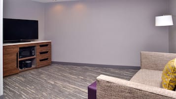 A modern living room with a flat-screen TV on a wooden cabinet, a sofa with a patterned cushion, and a lamp on the wall.