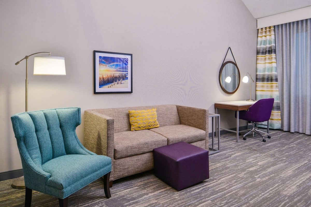 Una habitación de hotel con un sillón verde azulado, un sofá color crema con un cojín amarillo, un reposapiés morado, un escritorio con una silla morada, un cuadro en la pared y una lámpara de pie.