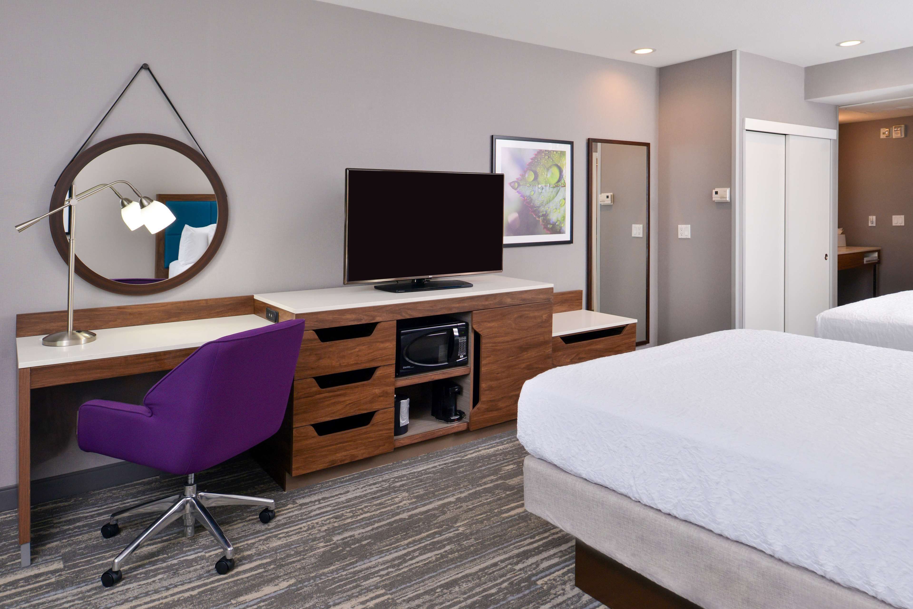 A hotel room with a wooden desk, a purple office chair, a flat-screen TV, a microwave, a bed, and a large mirror.