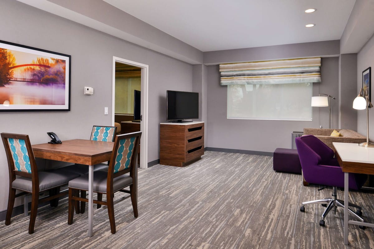 Habitación de hotel con una mesa de comedor, sillas, un televisor, un sofá y una ventana con persianas.