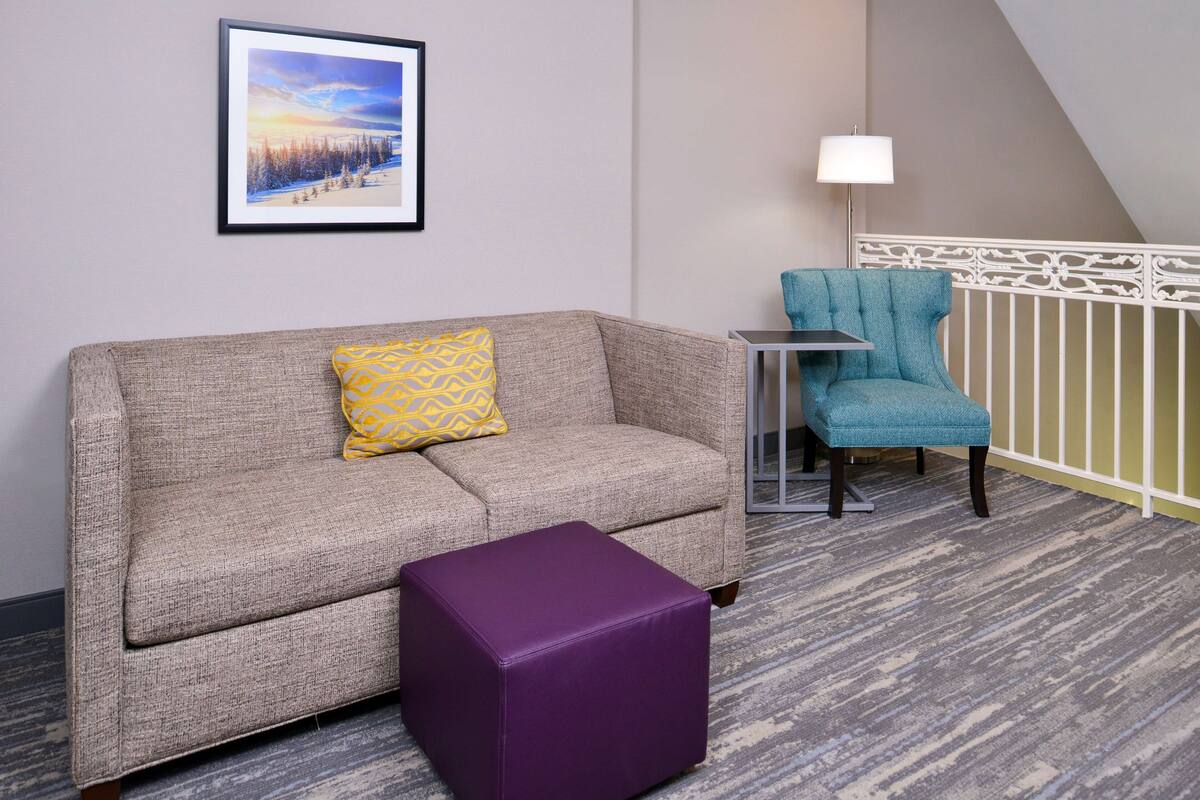 Una habitación de hotel con un sofá gris, un reposapiés morado, un sillón azul, un cuadro en la pared y una lámpara sobre una mesita de noche.