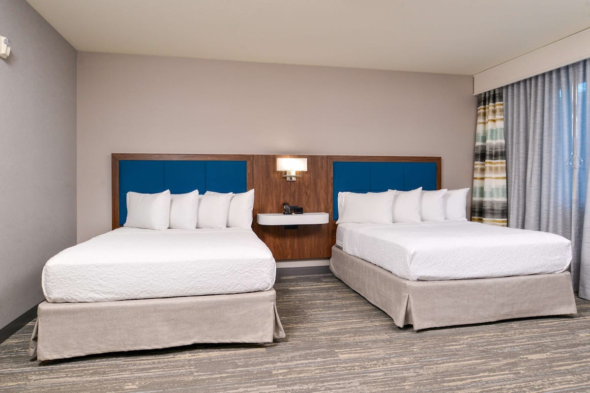 Habitación de hotel con dos camas, una mesita de noche pequeña y una ventana con cortinas.