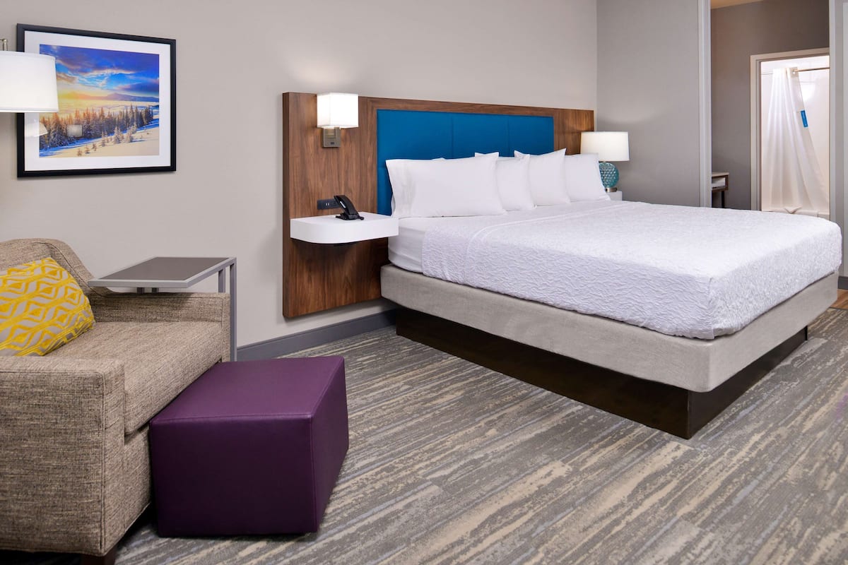 Habitación de hotel con una cama grande, una mesita de noche, un escritorio, una silla y un reposapiés morado.