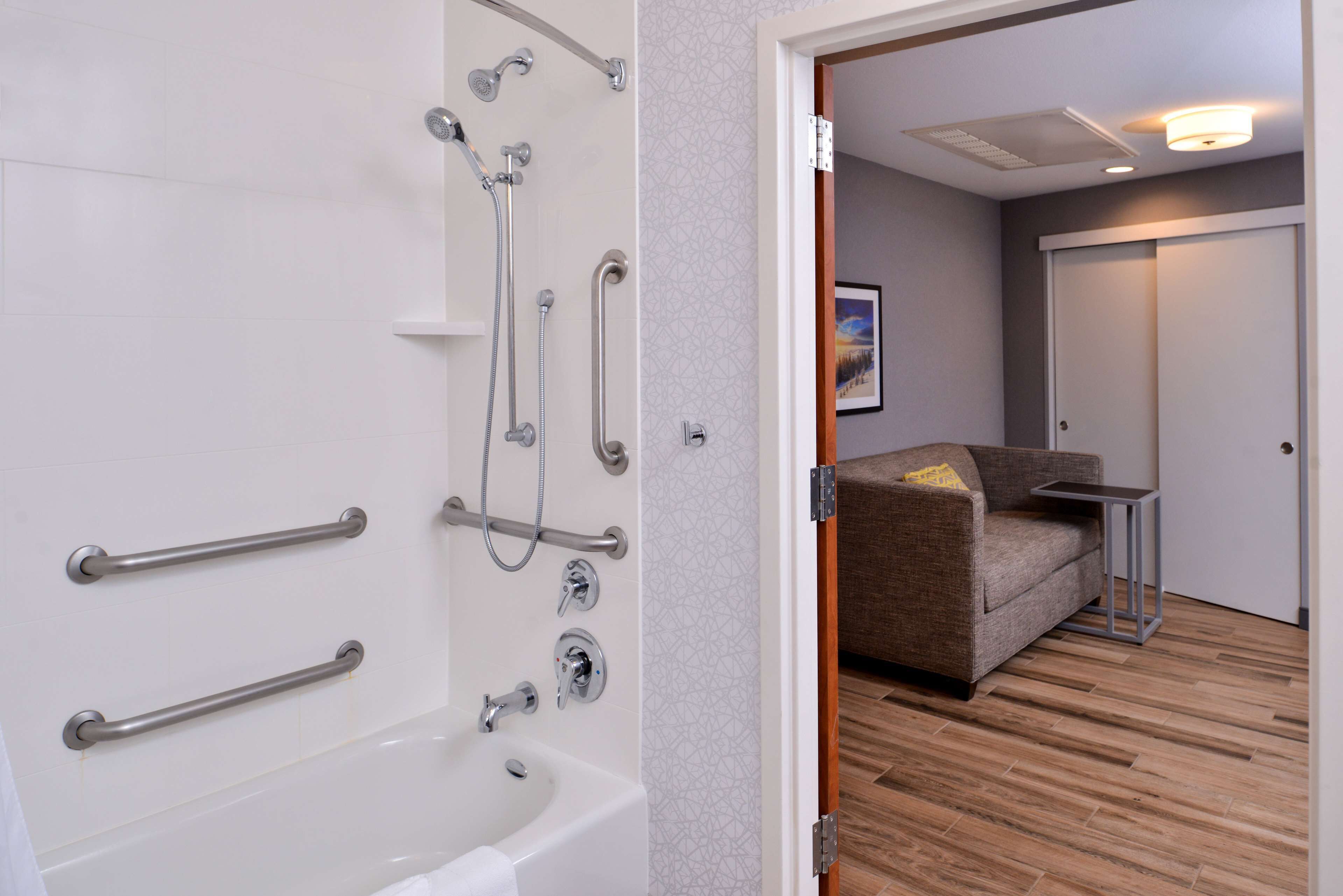 A bathroom with a bathtub, shower, and grab bars, a wooden floor, a gray sofa, and a small table.