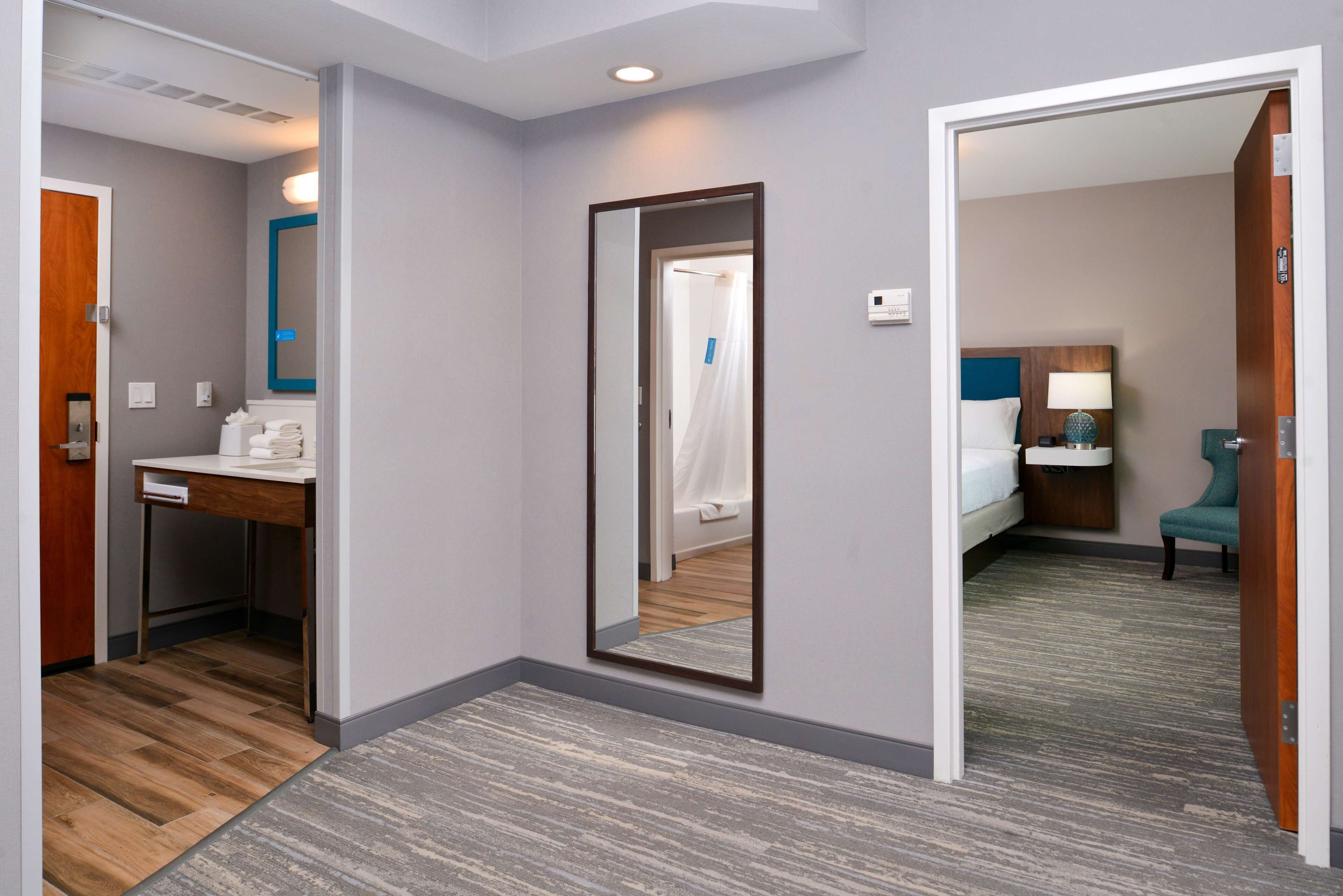 A hotel room with a desk, a mirror, a bed, a nightstand, and a chair.