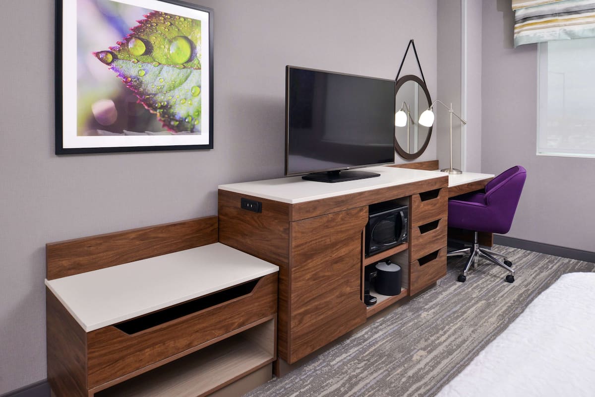 Una habitación de hotel moderna con un escritorio de madera, un televisor de pantalla plana, un espejo, una silla morada y una obra enmarcada en la pared.