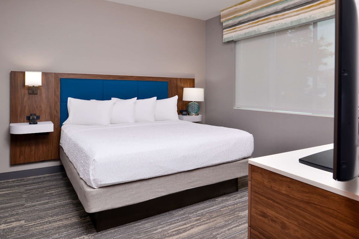 Habitación de hotel con una cama grande, una mesita de noche con lámpara, un televisor sobre un mueble y una ventana con persianas.