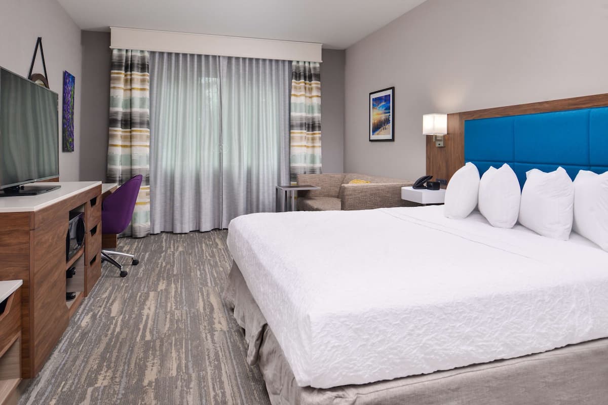 Habitación de hotel con una cama grande, un escritorio con computadora, una silla y una mesita de noche con lámpara.